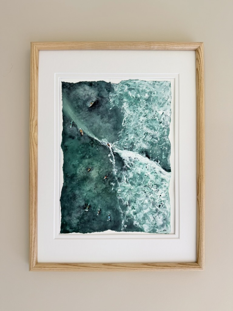 Framed aerial watercolor of surfers beside a breaking wave, matted in a light oak frame