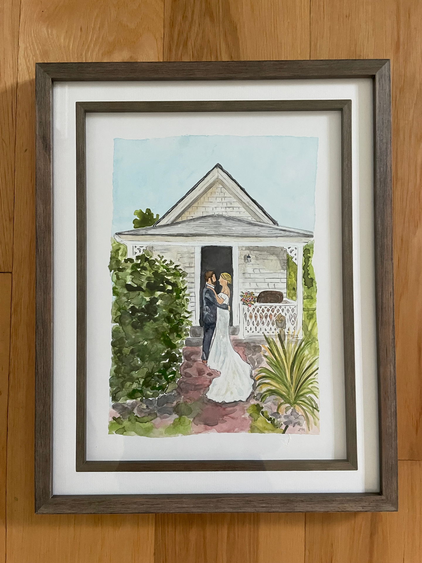 Framed watercolor of a bride and groom embracing in front of a coastal cottage