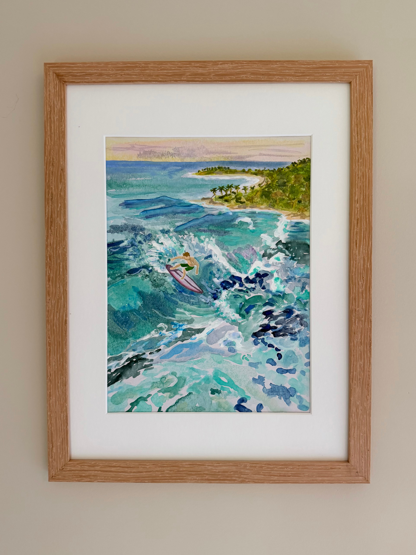 Framed watercolor of a lone surfer riding a turquoise wave near a tropical cove at sunset