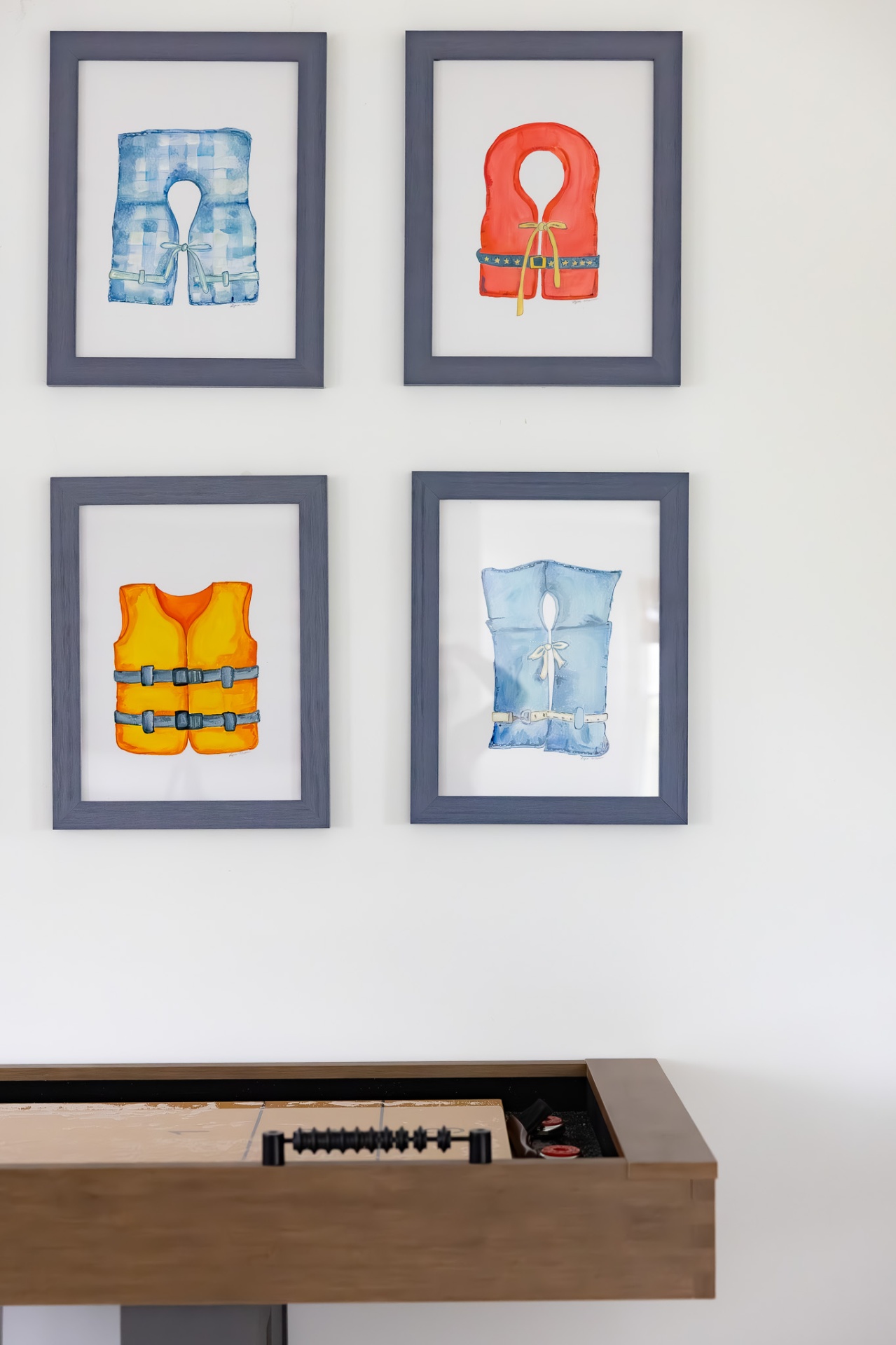 Four framed watercolor life vest paintings arranged in a two-by-two grid above a shuffleboard