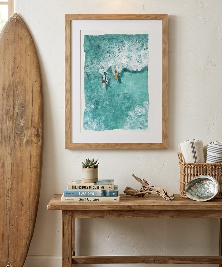 Framed watercolor of two surfers paddling into turquoise surf, styled above a rustic console with books and a surfboard