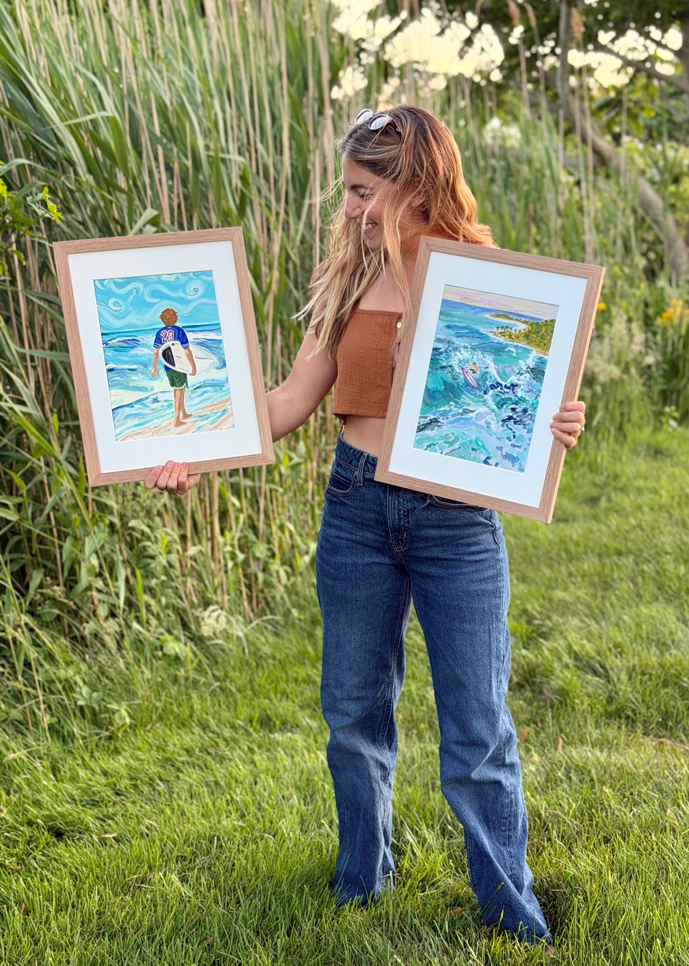 Lynn standing in the reeds holding two framed watercolor paintings of surfers