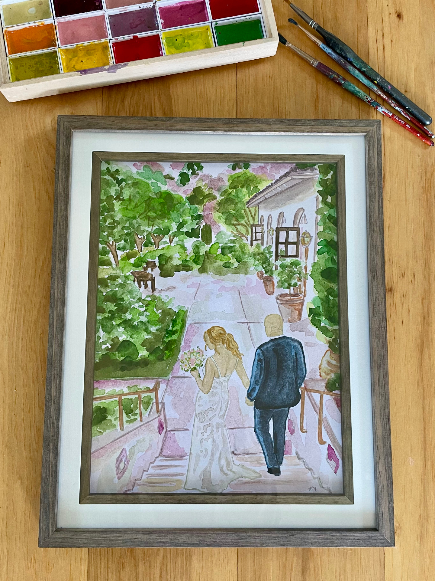 Framed watercolor of a bride and groom walking through a lush garden courtyard