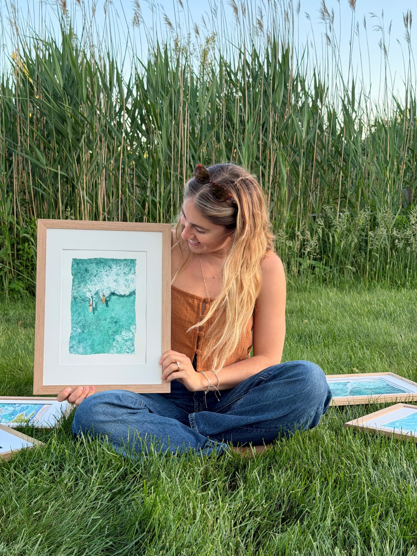 Lynn sitting in the grass holding a framed aerial watercolor of two surfers