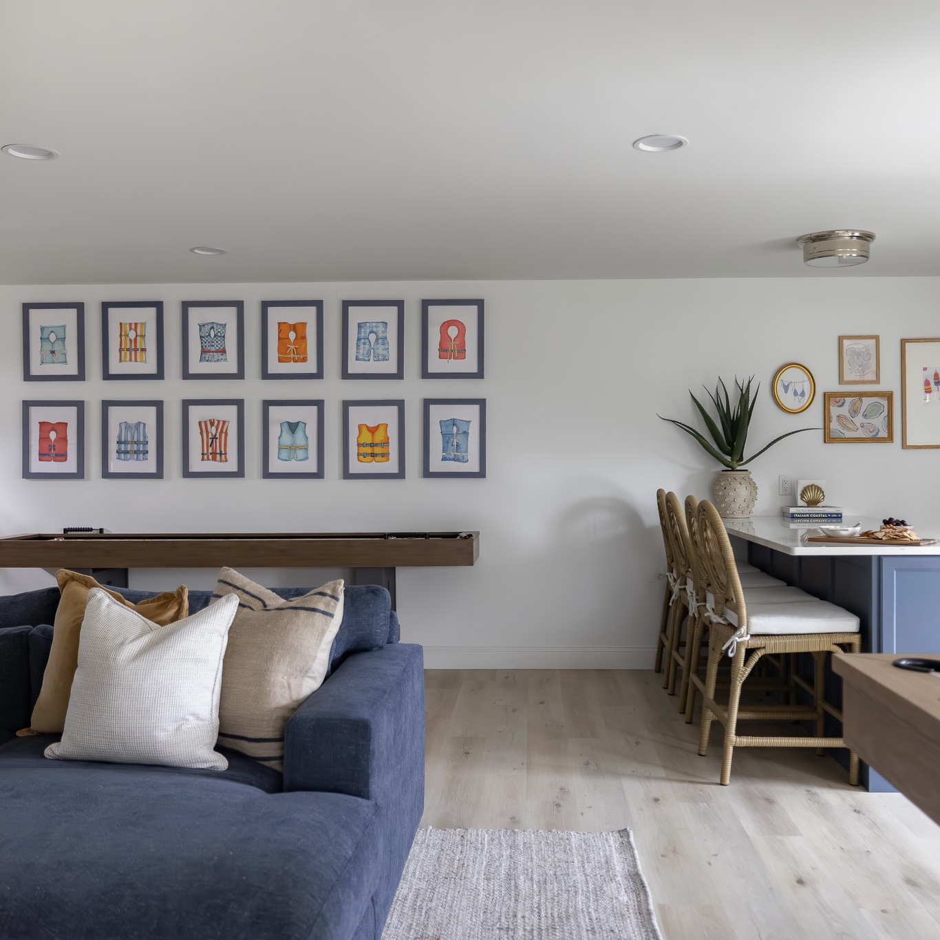 Two rows of framed watercolor life vest paintings hung above a shuffleboard table in a coastal lounge