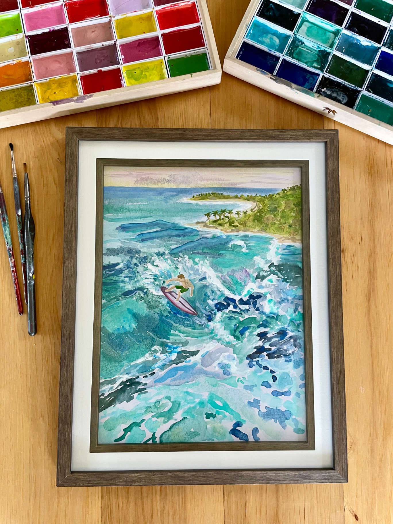 Framed aerial watercolor of a surfer riding a turquoise wave near a tropical shore