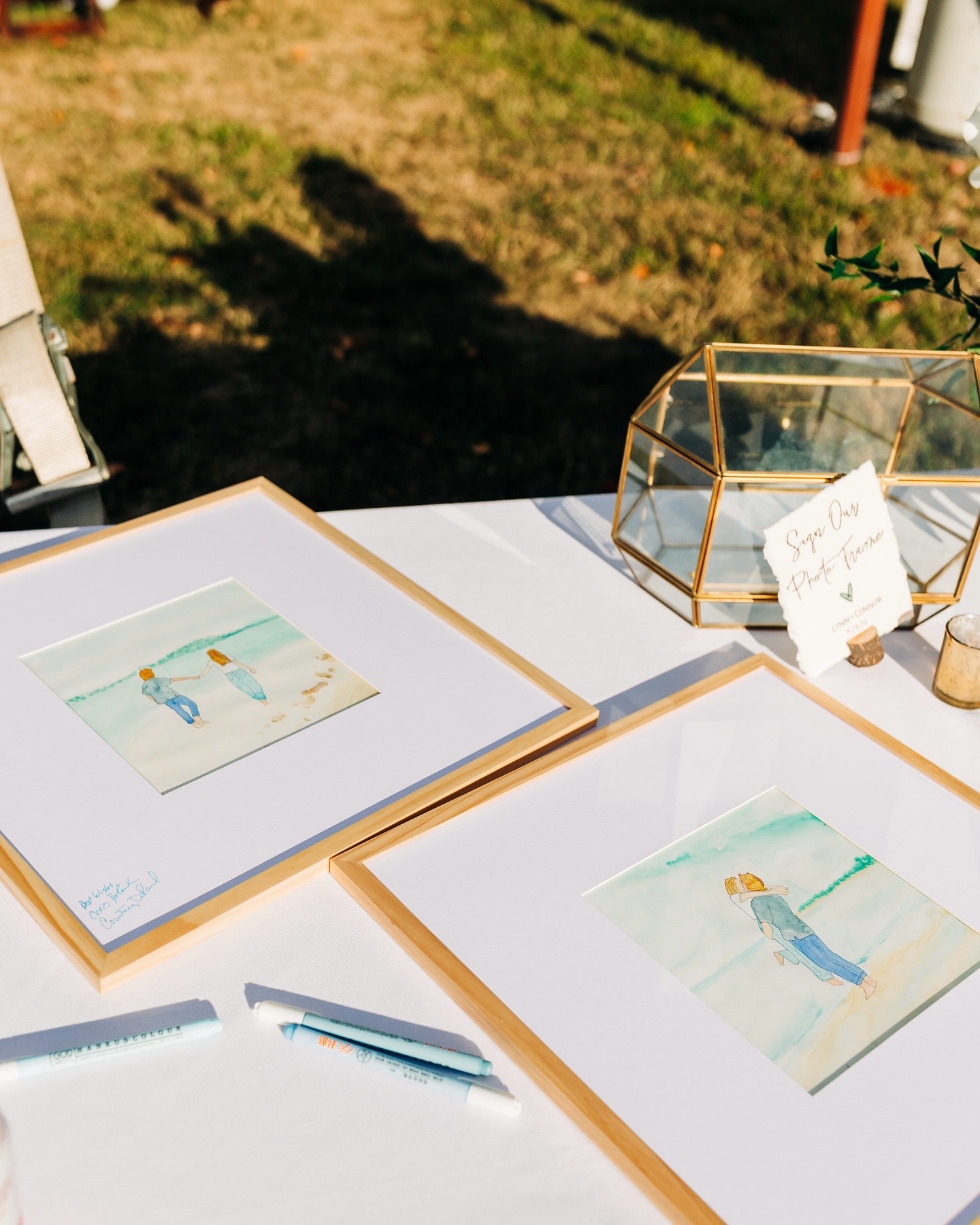 Two framed watercolor wedding portraits displayed on a table beside a sign our photo frame card at an outdoor wedding
