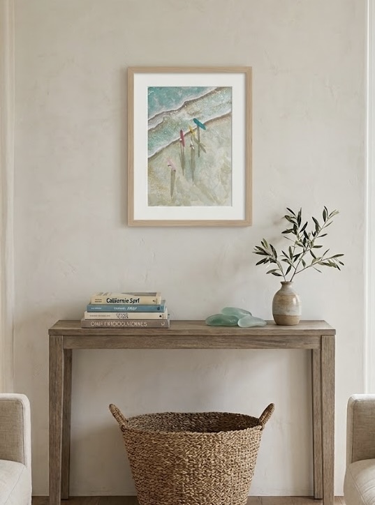 Framed aerial watercolor of colorful surfboards on a sandy shore, hung above a console with surf books